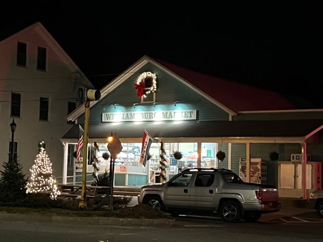 a small market building decorated for the holidays at night, with a lit christmas tree outside and a pickup truck parked in front.