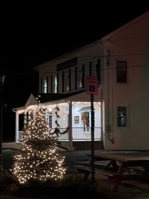 a christmas tree with white lights stands near a building decorated with holiday wreaths and string lights at night.