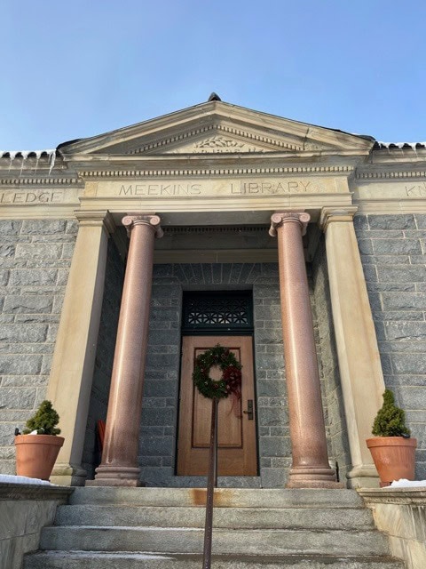 stone entrance of meekins library with two pink columns, a wooden door decorated with a wreath, and potted plants on either side.