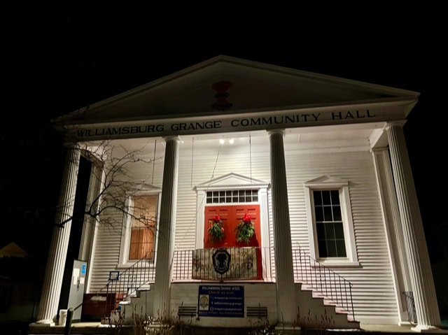 williamsburg grange community hall at night, illuminated, with white pillars, a red double door, wreaths, and signs near the entrance.