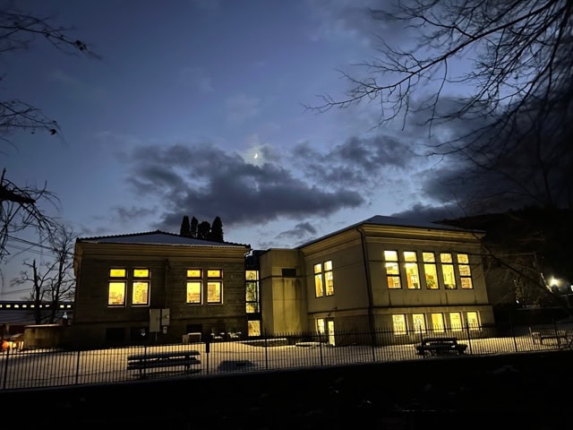 a two story building with large lit windows stands in a snowy landscape at dusk, with dark clouds and faint moon visible in the sky.