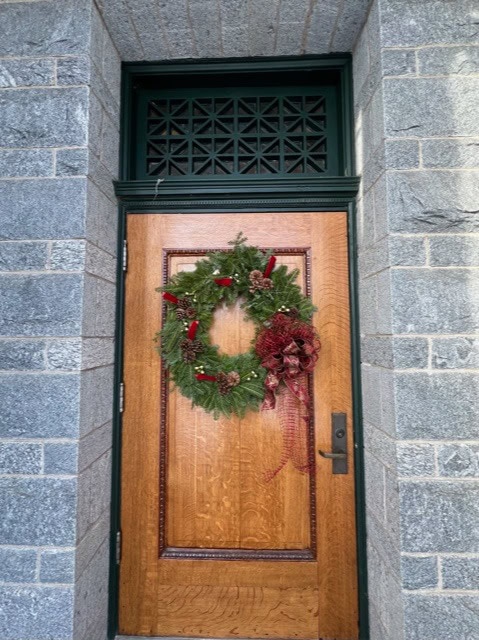 a wooden door set in a gray stone wall, decorated with a holiday wreath made of greenery, pine cones, red accents, and a red ribbon.