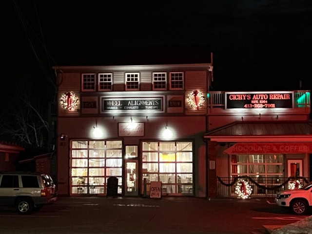 a two story building at night with illuminated signs for wheel alignments, chocolates and coffee, and an auto repair shop. a white suv is parked in front. holiday wreaths decorate the facade.