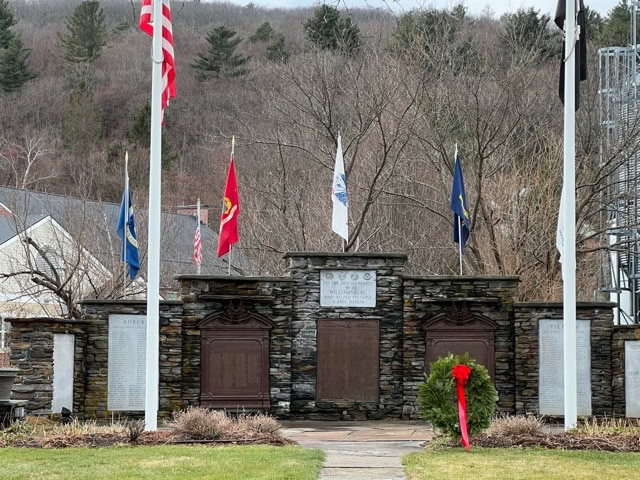 a stone veterans memorial with plaques, inscribed names, five flagpoles displaying flags, and a red wreath in front, set against trees and a cloudy sky.