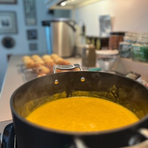 a pot of orange colored soup simmers on a stove, with muffins and various kitchen items blurred in the background.