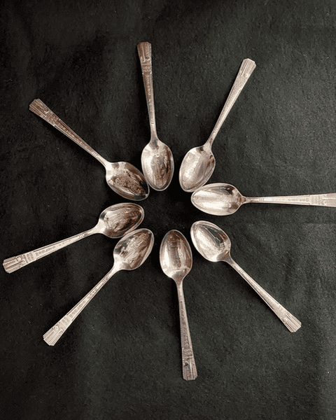 eight metal teaspoons are arranged in a circular pattern on a black surface, with their handles pointing outward and bowls meeting in the center.