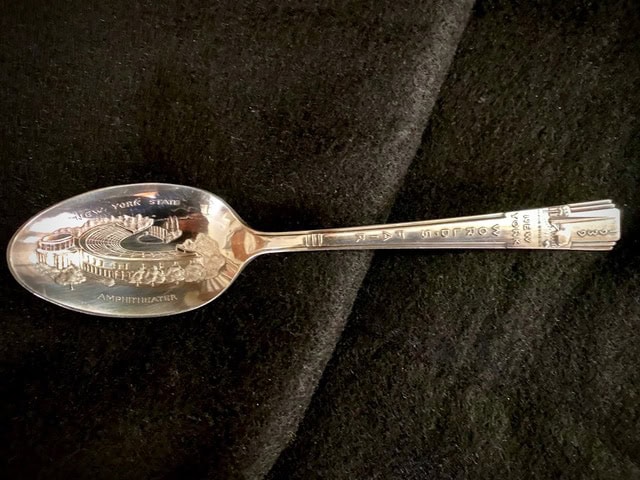 a commemorative silver spoon featuring an engraved image of the new york state fair amphitheater on a black fabric background.