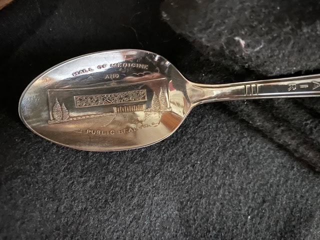 a silver spoon with an engraved image and the text "school of medicine and public health" on its bowl, resting on a dark fabric surface.