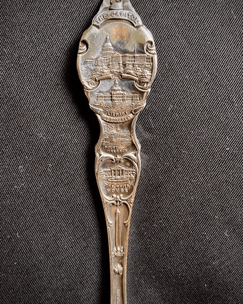 a souvenir spoon with embossed images and labels of the capitol, library of congress, washington monument, and the white house on a dark textured background.