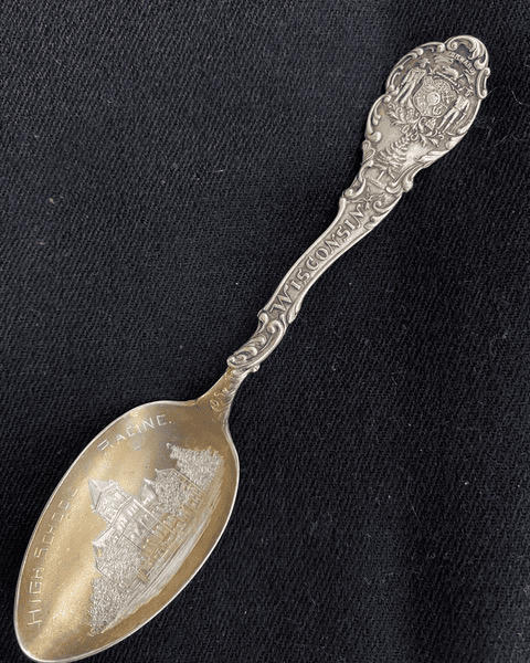 antique souvenir spoon with engravings of "wisconsin" on the handle and a detailed building labeled "high school racine" in the bowl, set against a black background.