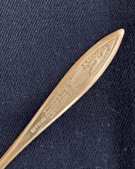 a brass souvenir spoon with an engraving reading "a century of progress, official, 1933 chicago" on a textured dark fabric background.