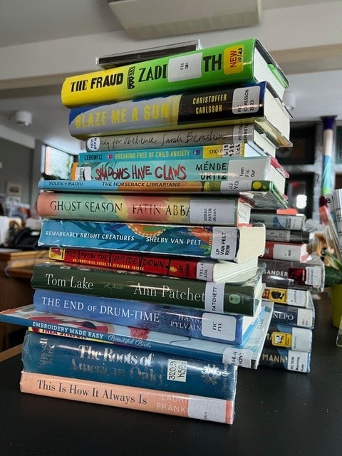 A tall stack of assorted hardcover and paperback library books sits on a table in a home setting, with visible title labels and barcodes.