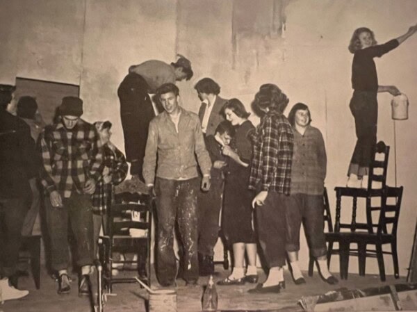 a group of people, mostly young adults, stand and work together in a room. some are on chairs painting the wall, while others gather and watch.