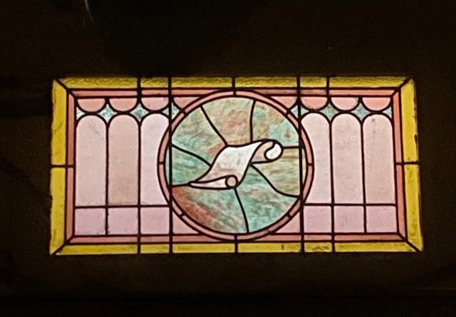 rectangular stained glass window with a central circular panel showing a rolled scroll, surrounded by pastel pink, yellow, and green geometric patterns.