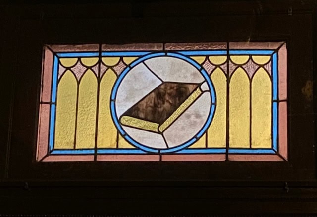 stained glass window featuring a geometric design with a central book shape surrounded by yellow and blue patterns.