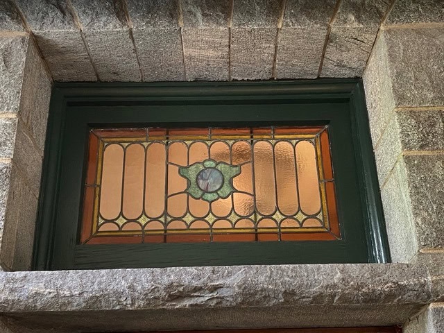 rectangular stained glass window with a green, brown, and amber geometric pattern, set in a stone frame above a doorway.