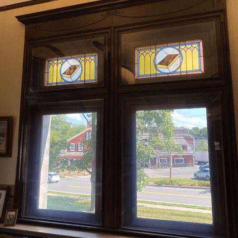 two tall windows with stained glass panels at the top, showing a street, trees, and buildings outside on a sunny day.