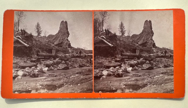 stereoscopic image of a rocky landscape with sparse trees, uneven terrain, and scattered logs or debris, bordered by an orange frame.