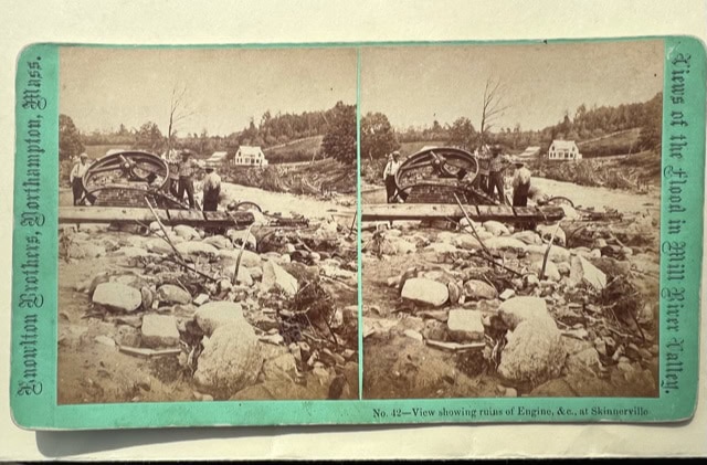 two men stand among debris and the ruins of machinery in a rocky landscape, with houses and trees in the background; photo labeled as mill river valley flood aftermath.