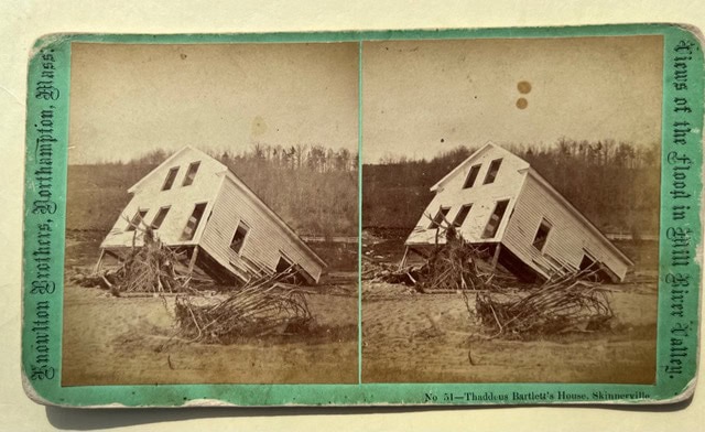 a house leaning heavily to one side sits among flood debris, with barren trees and a river in the background.