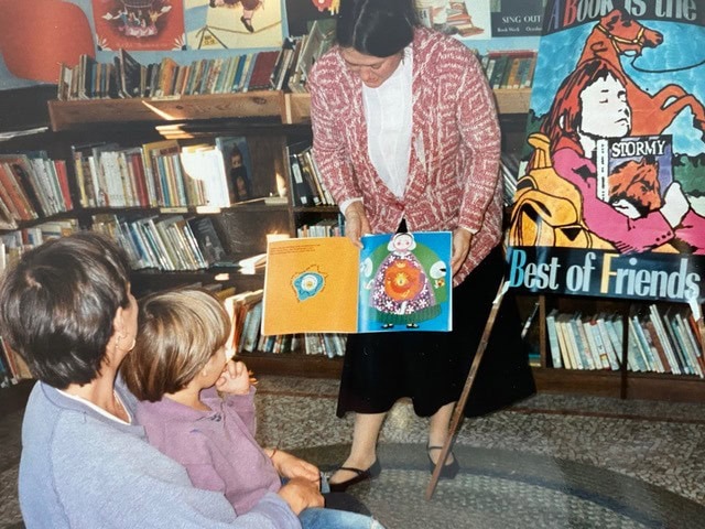 a woman reads a children's book to two kids in a library, with bookshelves and a "best of friends" poster visible in the background.