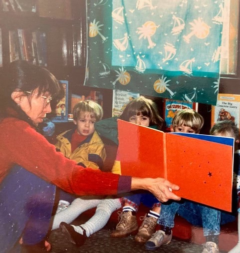 an adult reads a large picture book to a group of young children sitting on the floor in a library or classroom.