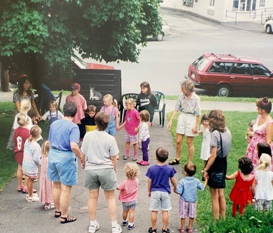 a group of adults and children stand in a circle on a driveway and grass area, participating in an outdoor activity near parked cars and trees.