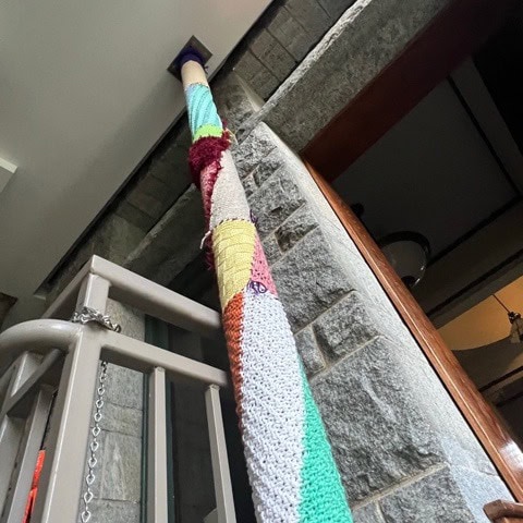 a vertical pole wrapped in colorful yarn stands next to a stone wall and a metal railing inside a building.