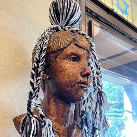a bronze bust of a child is adorned with a black and white yarn pom pom hat with long tassels, displayed indoors near a window.