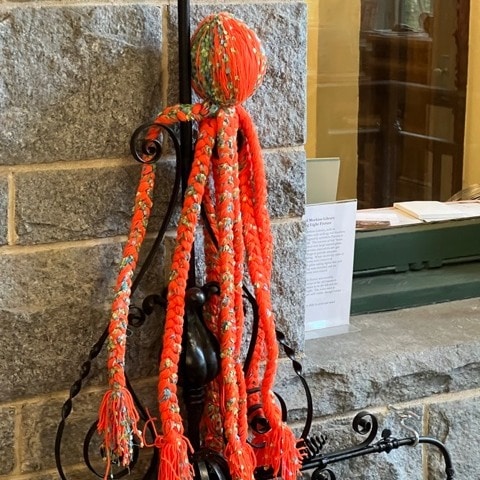 a ball of multicolored yarn with several long, thick, braided strands hangs from a decorative black metal stand against a stone wall.