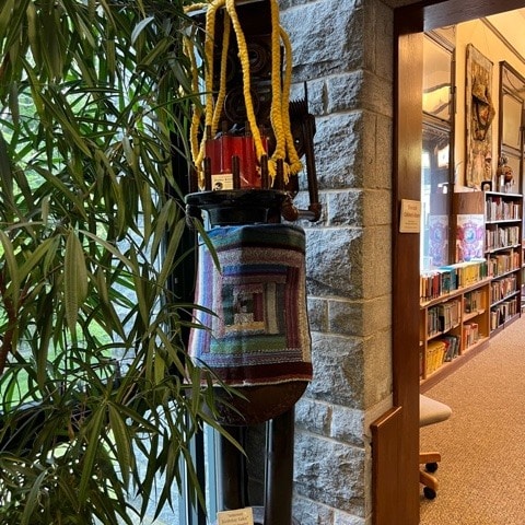 a colorful woven bag hangs from a black stand near a stone wall and a large plant, with bookshelves visible in the background.