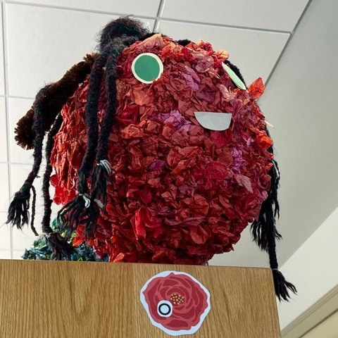a large red, textured paper sculpture with green eyes, black yarn hair, and a white smile sits atop a wooden shelf next to a flower sticker.