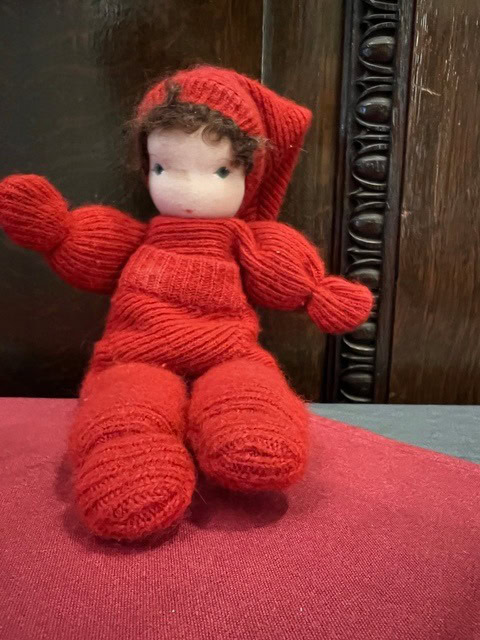 a handmade cloth doll with curly brown hair, blue eyes, and a red knitted outfit sits on a red surface in front of a dark wooden background.