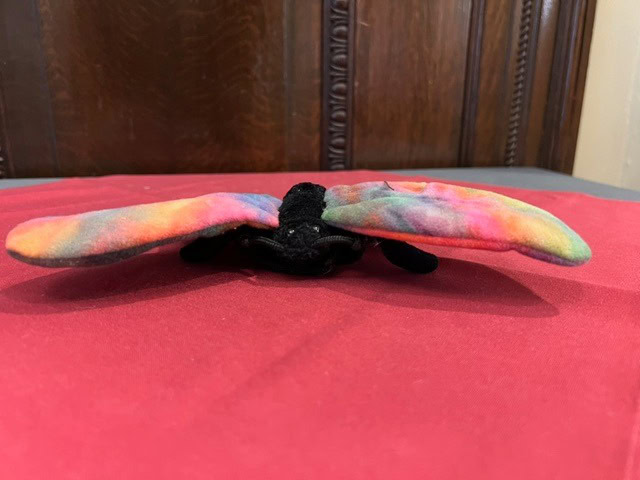 a plush toy with brightly colored, tie dye patterned wings sits on a red surface in front of a wooden background.