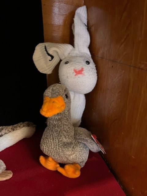 a white stuffed rabbit and a gray stuffed duck with orange beak and feet are positioned against a wooden wall on a red surface.