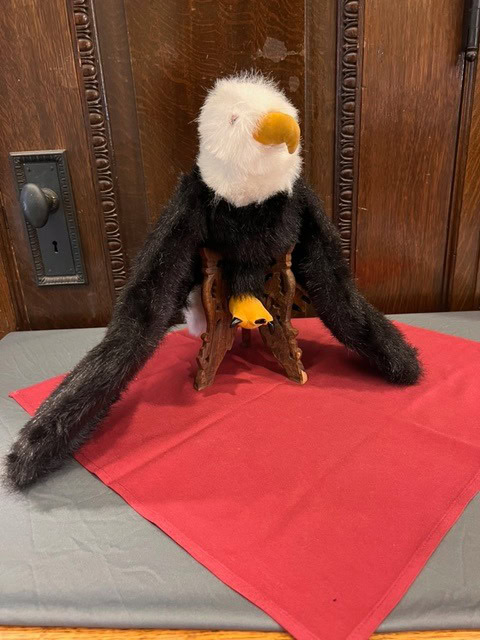 a plush bald eagle with long black wings is seated on a wooden stand, placed on a red cloth atop a bench in front of a wooden wall.