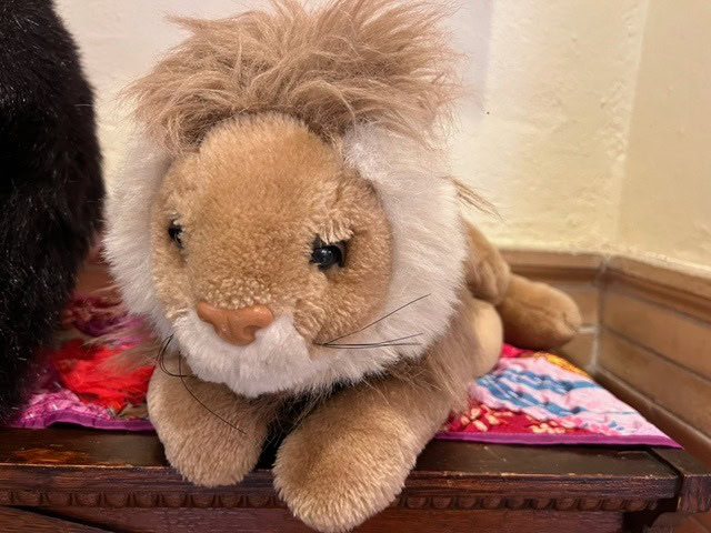 a brown and white plush lion toy lies on a colorful quilt, placed on a wooden surface against a cream colored wall.
