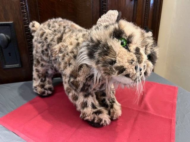 a plush toy cat with spotted fur and green eyes stands on a red cloth on a gray surface, in front of a wooden background.