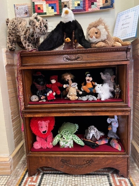 a wooden bookshelf displays various stuffed animals and dolls, including a lion, eagle, frog, pink bear, and elephant, arranged on three shelves.
