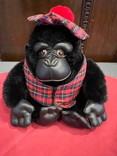 a plush gorilla toy wearing a red plaid vest and matching hat sits on a red surface in front of a wooden background.