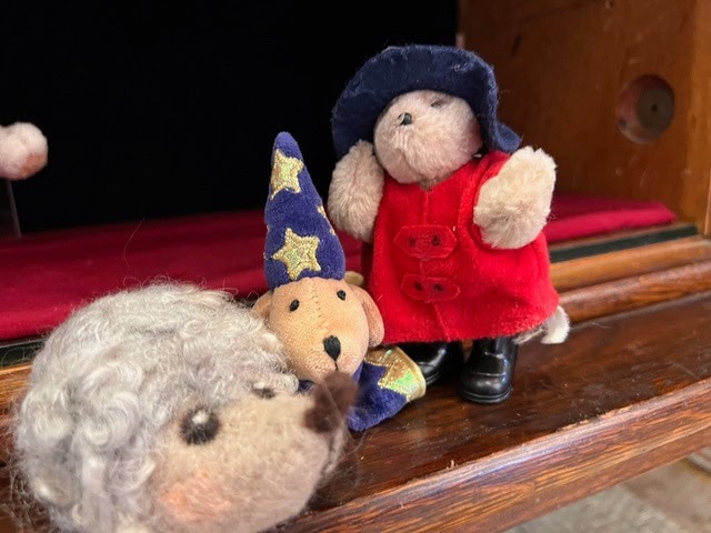 three stuffed animal toys, including a bear in a red coat and hat, a wizard bear with a starry hat, and a gray sheep, are placed on a wooden shelf.