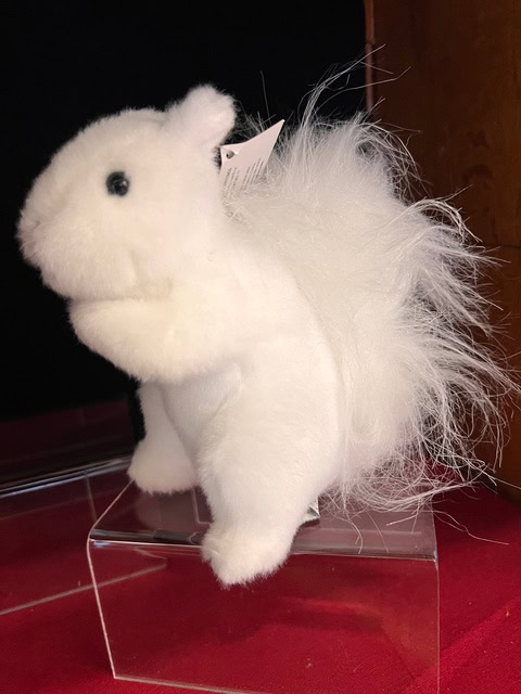 a white plush squirrel with a fluffy tail is displayed on a clear plastic stand against a dark background.