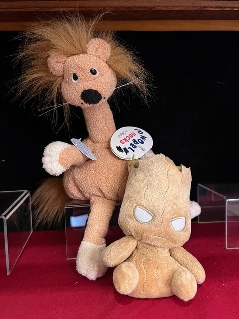 two plush toys are displayed on a red surface: a tan lion with a “roar! press me!” button and a small, light brown groot figure with white eyes in front.