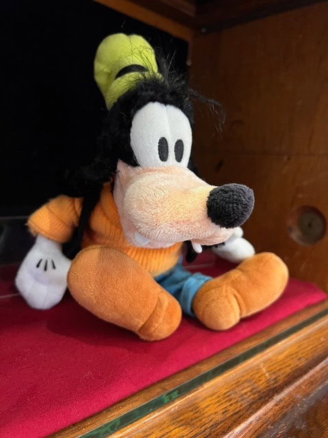plush toy of goofy, wearing an orange shirt, blue pants, and green hat, sitting on a red surface against a wooden background.