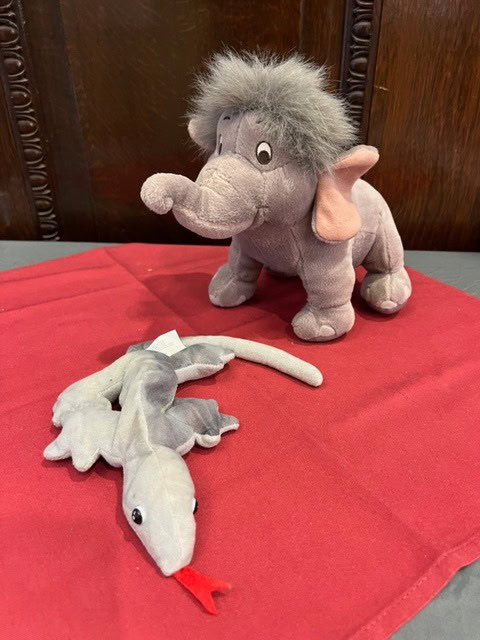 a gray plush elephant and a gray plush lizard with a red tongue are displayed on a red cloth with a wooden background.