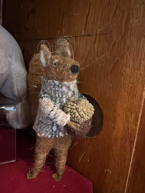 a small, handmade squirrel figurine made of textured materials holds a pinecone and stands against a wooden background.