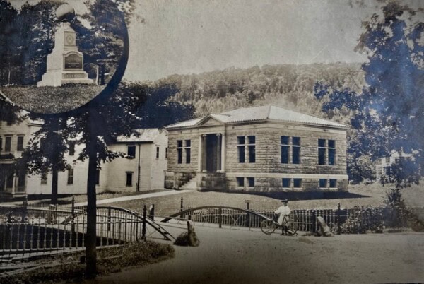 a sepia tone photo of a stone library building with large windows, a small bridge in the foreground, and a monument inset in the top left corner. trees and houses surround the scene.