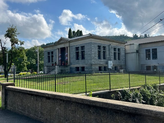 a large, gray stone building with columns and banners stands behind a metal fence on a sunny day, with trees and clouds in the background.