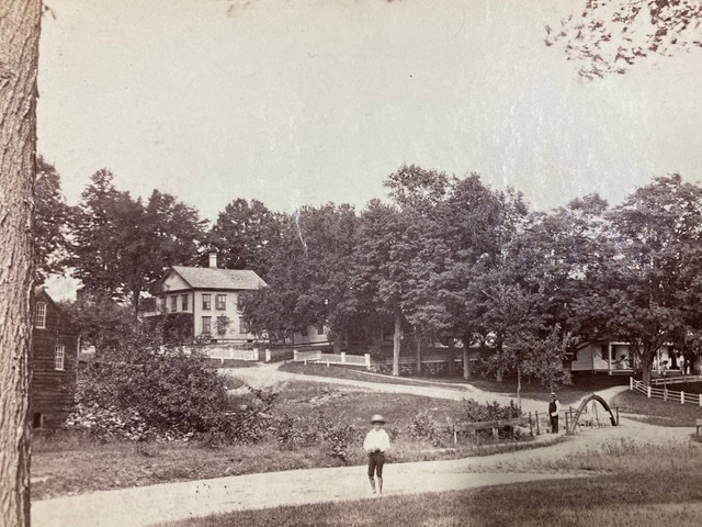 a young boy stands on a path in front of a small bridge, with trees and two large houses in the background.