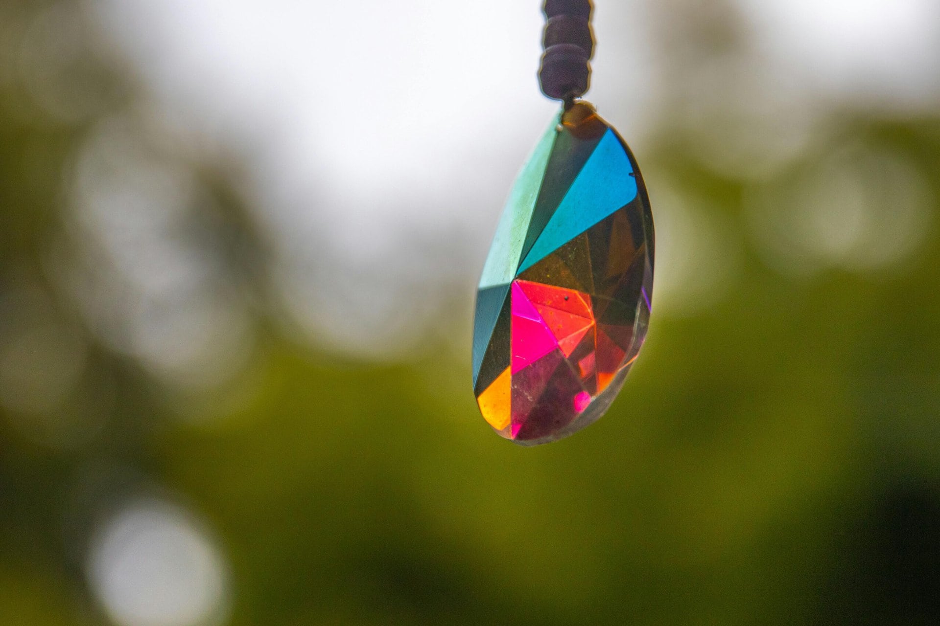 a close up of a faceted, multicolored crystal pendant hanging outdoors with a blurred green background.